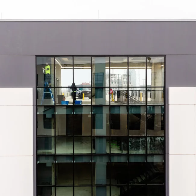 Modern parking garage in Mobile, AL with a tall glass atrium and white facade beside an active construction site, cranes, and a striped roadway in daylight.
