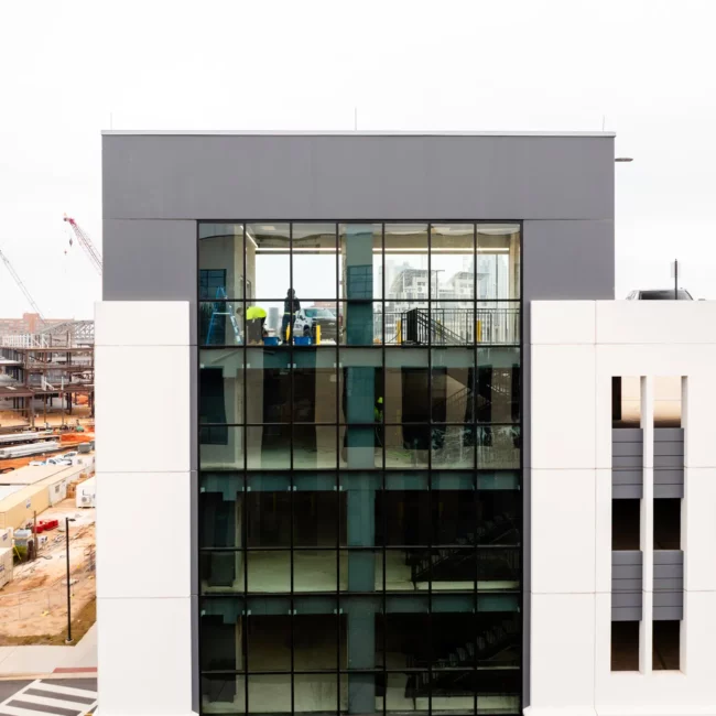 Modern parking garage in Mobile, AL with a tall glass atrium and white facade beside an active construction site, cranes, and a striped roadway in daylight.