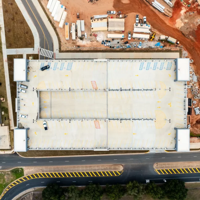 Aerial view of a large warehouse under construction beside roads, striped crossings, parked vehicles, and red-earth construction materials.