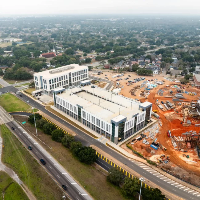 post construction cleaning job site in mobile alabama Aerial view of a modern white building beside a busy highway, with an active construction site and sprawling suburban neighborhood beyond.