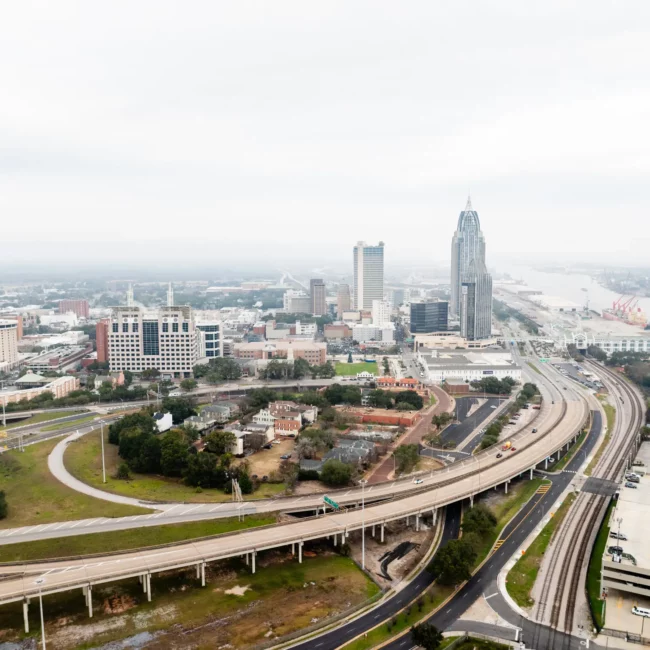 Aerial view of Mobile, Alabama, with the tall RSA Battle House Tower, neighboring buildings, roads, and distant coastline under a hazy sky.