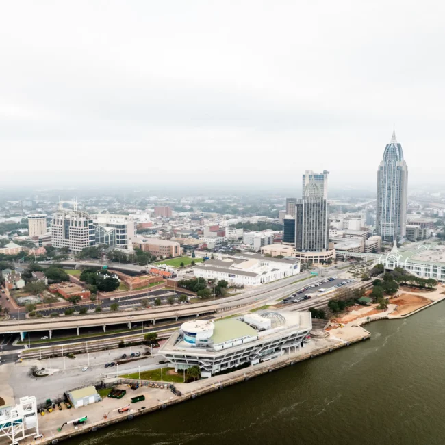 Aerial view of Mobile, Alabama, with the tall RSA Battle House Tower, neighboring buildings, roads, and distant coastline under a hazy sky.