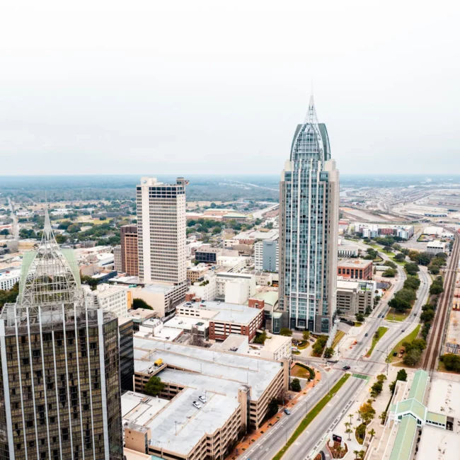 Aerial view of Mobile, Alabama, with the tall RSA Battle House Tower, neighboring buildings, roads, and distant coastline under a hazy sky.