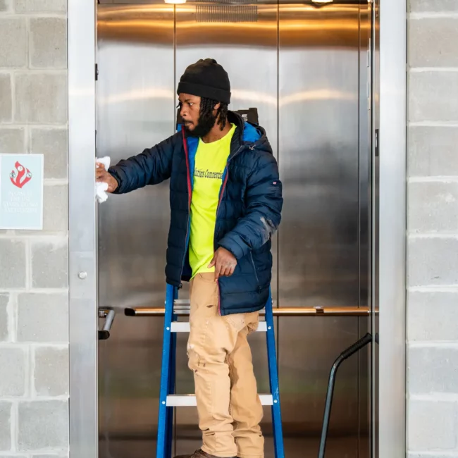 Cleaning worker in a blue jacket and tan pants stands on a step stool inside an open elevator, reaching up; yellow mop bucket rests below.