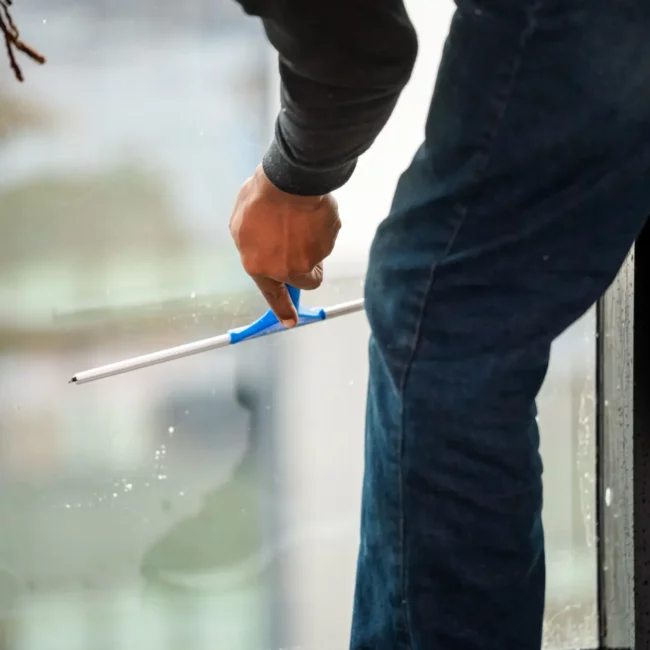 Window cleaner using a blue squeegee on a glass pane, viewed close-up; workwear and focused movement convey professional maintenance.