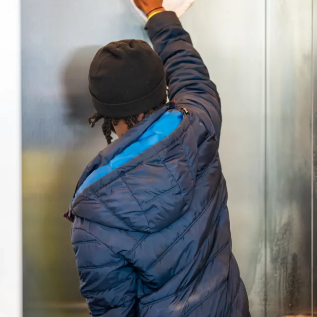 A person in a navy puffer jacket and black cap wipes a metal elevator wall with a white cloth, conveying careful indoor cleaning and upkeep.