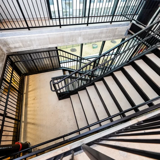 Looking down an industrial concrete stairwell, black metal railings zigzag between levels, creating a bright, geometric, dizzying view.