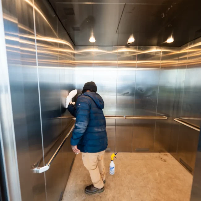 Cleaner in a blue jacket mops the floor of a reflective stainless-steel elevator, with a spray bottle nearby and warm ceiling lights glowing.