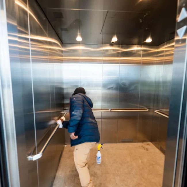Cleaner in a blue jacket mops the floor of a reflective stainless-steel elevator, with a spray bottle nearby and warm ceiling lights glowing.