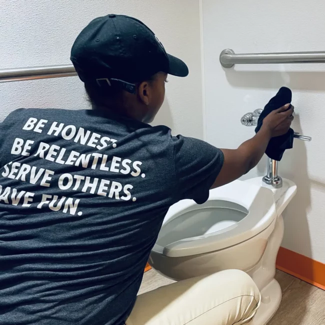 Person in a black cap wipes a restroom toilet while wearing a shirt reading, “Be honest. Be relentless. Serve others. Have fun.”